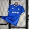 Camiseta oficial de 'El Barrio' de la King League en color azul, con logo de InfoJobs en el centro y detalles en dorado en el cuello y las mangas, colgada en una rejilla negra.