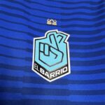 Primer plano del escudo del equipo 'El Barrio' de la King League en la camiseta oficial, sobre fondo azul con líneas horizontales, y detalle de dos coronas doradas encima del escudo.